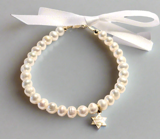 A white bracelet made of pearls and 14 KT beads with a gold-colored Star of David charm, presented against a white background with a white ribbon and a gold ribbon.